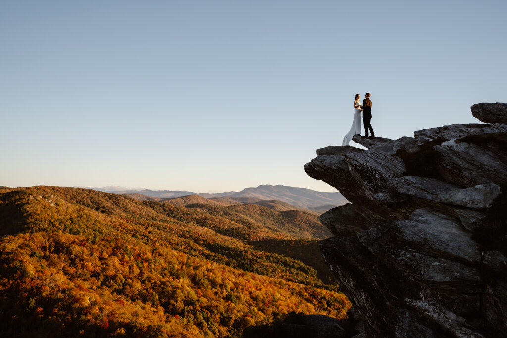 Couple standing on a cliff edge with fall foliage and mountains beneath them