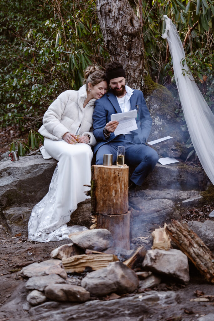 Couple in wedding attire sitting together reading letters from their families next to a campfire